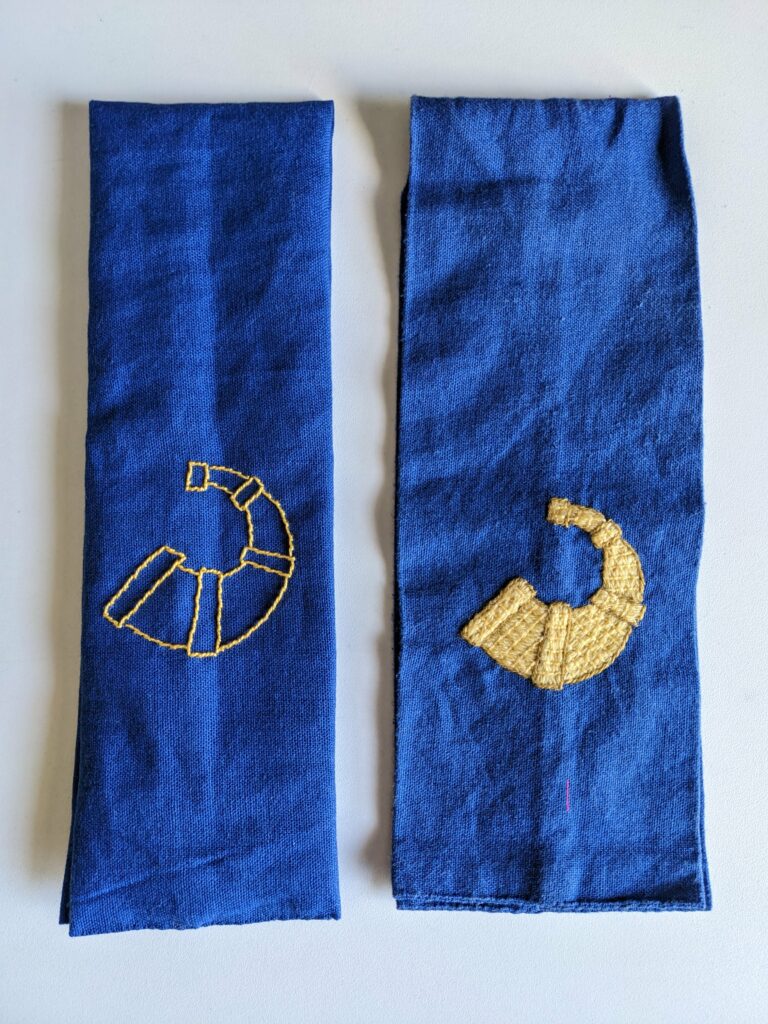Two Hartshorn-Dale favors, strips of blue fabric with yellow hunting horns embroidered on. One horn is outlined and the other is filled in.